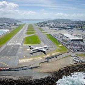 A drone shot of the runway at Wellington International Airport with a plane on the runway, a parking lot filled with cars located at Stewart Duff Drive, Rongotai with Hataitai, Evans Bay and Miramar seen in the background.