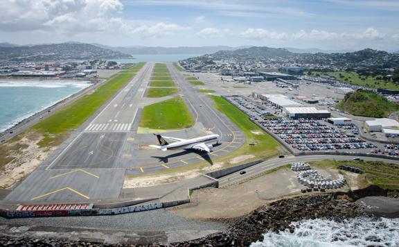 A drone shot of the runway at Wellington International Airport with a plane on the runway, a parking lot filled with cars located at Stewart Duff Drive, Rongotai with Hataitai, Evans Bay and Miramar seen in the background. 