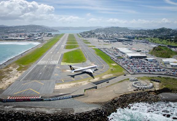 A drone shot of the runway at Wellington International Airport with a plane on the runway, a parking lot filled with cars located at Stewart Duff Drive, Rongotai with Hataitai, Evans Bay and Miramar seen in the background. 