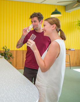 Two smiling people try craft beers at Parrotdog Brewery in Lyall Bay, Wellington. The yellow-tiled wall behind them brightens the space and The bartender behind the counter is holding the next two beer samples.