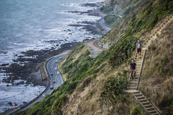 Two people walking down wooden steps on the Escarpment Track. The coastal road in the background has many cars on it, and nearby are large rocky outcrops on the shoreline.