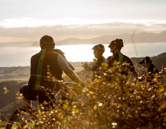 Four cyclists stop to rest by a bush in the early morning sunlight on Red Take Track at Whareroa Farm.