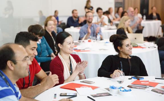 In the foreground sits 4 people listening and clapping to someone in the distance, they are sat at a table with notepads in front. In the background are more people blurred out.