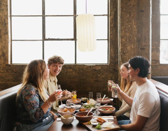 Four young adults enjoy drinks and food in a booth table under a large window inside Chow, on Tory Street.