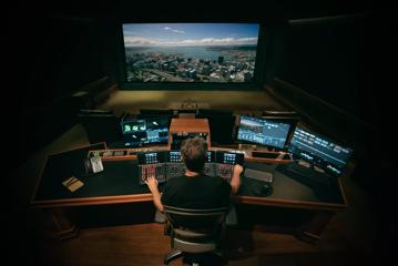 A person working on post-production for a film at a large desk with a sound board, three computer monitors and a projection screen.