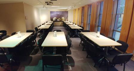 A conference room set up with nine tables with eight chairs, a water jug and glasses at each of them.