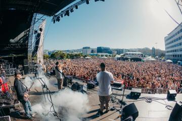 Four musicians perform on stage to a packed audience at an outdoor music festival.