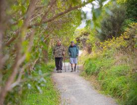 Two people using hiking sticks walk along a gravel path in a forest.