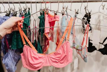 A hand with red painted nails holding a pink bra on a wire hanger.