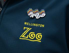 A person's left shoulder, wearing a navy blue zipped jacket, with the Wellington Zoo logo embroidered in yellow and a rainbow pin that says "proud" fixed above it.