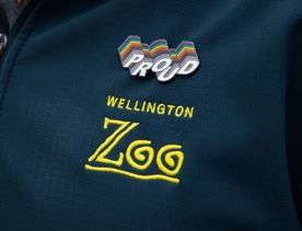 A person's left shoulder, wearing a navy blue zipped jacket, with the Wellington Zoo logo embroidered in yellow and a rainbow pin that says "proud" fixed above it.