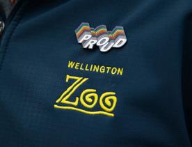 A person's left shoulder, wearing a navy blue zipped jacket, with the Wellington Zoo logo embroidered in yellow and a rainbow pin that says "proud" fixed above it. 