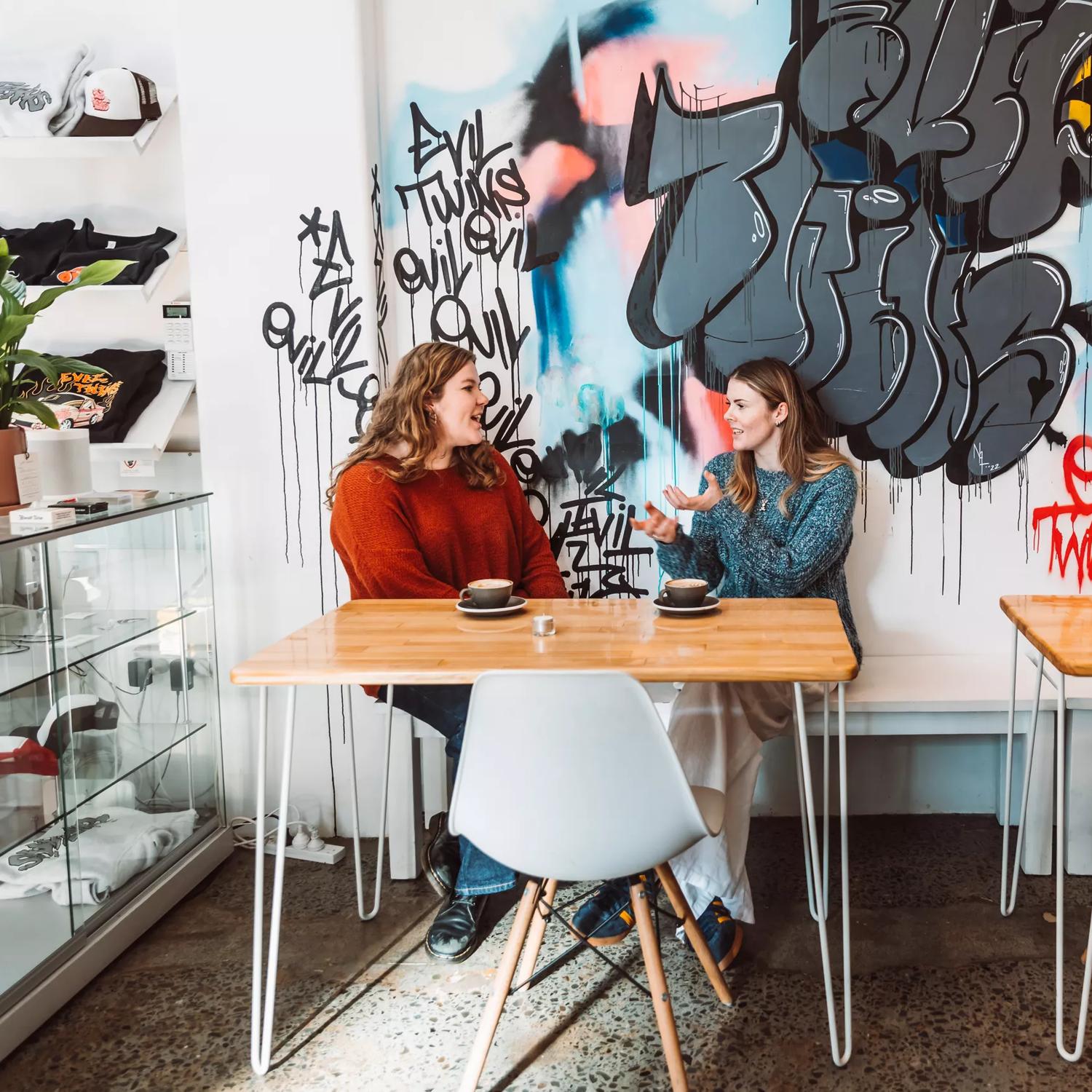 Two people sit at a table by a white wall covered in graffiti chatting over coffee inside Evil Twins  Coffee, a café in Te Aro Wellington.