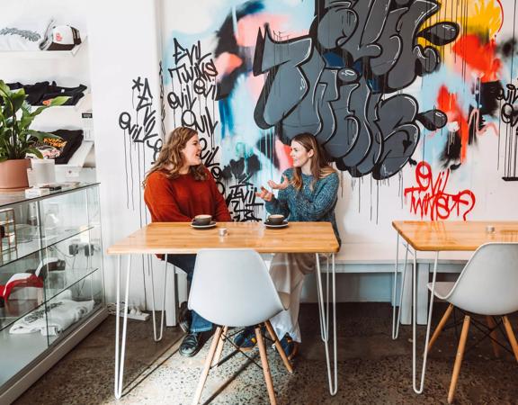 Two people sit at a table by a white wall covered in graffiti chatting over coffee inside Evil Twins  Coffee, a café in Te Aro Wellington.