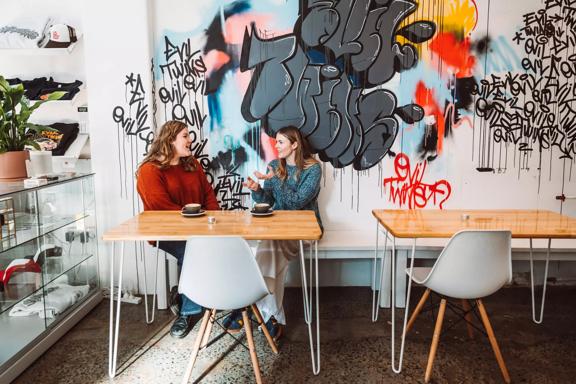 Two people sit at a table by a white wall covered in graffiti chatting over coffee inside Evil Twins  Coffee, a café in Te Aro Wellington.