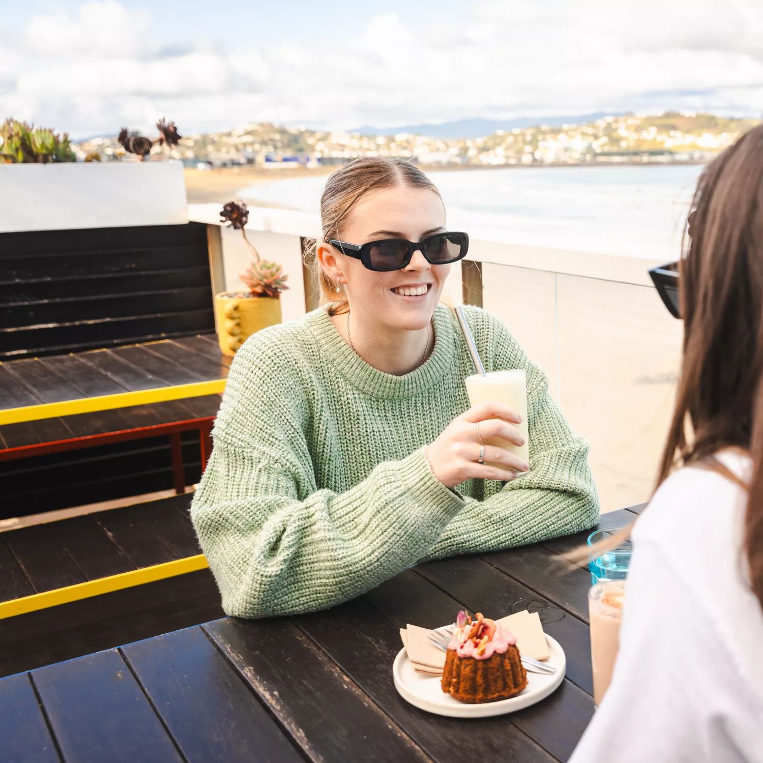 Two friends sit on a patio and enjoy milkshakes while overlooking the beach.