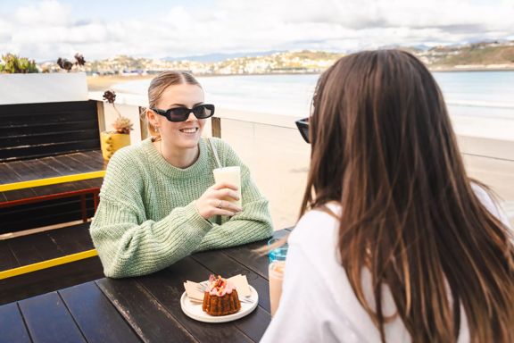 Two friends sit on a patio and enjoy milkshakes while overlooking the beach.