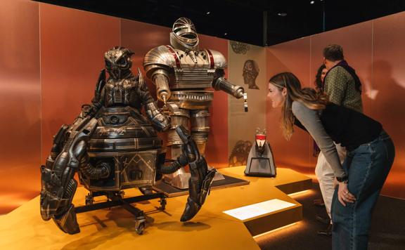 A person crouching over and looking at metal robots that are part of an exhibit. The room has red walls and orange flooring.