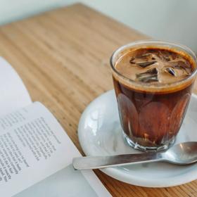 A small glass of iced coffee is on a white saucer with a small silver spoon on wooden table surface next to an opened magazine.