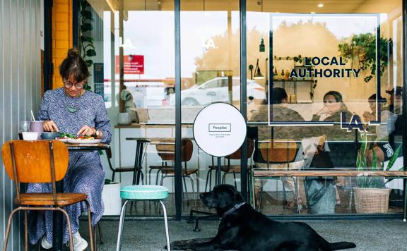 A person sitting outside Local Authority in Porirua eating food. A black labrodor dog sits at their feet.