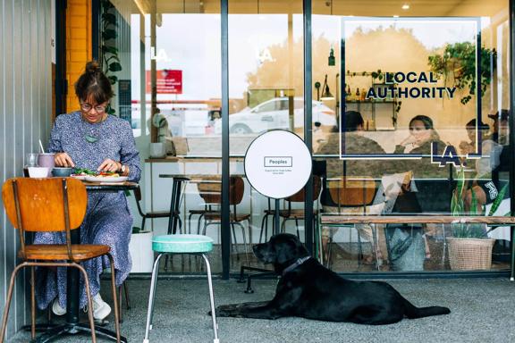 A person sitting outside Local Authority in Porirua eating food. A black labrodor dog sits at their feet.