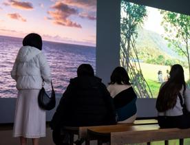 Four people, one stands and three are seated on a bench, observing photographs of natural landscapes projected onto the back wall. 