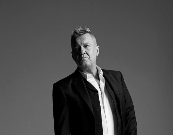 Jimmy Barnes, a Scottish-born Australian rock singer, wears a suit and poses in front of a solid colour backdrop.