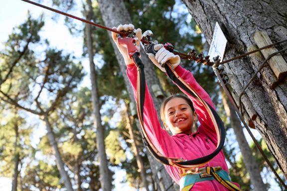 A child wearing a pink sweater and white gloves looks down smiling from a tree top trek at Adrenalin Forest in Aotea, Porirua, New Zealand.