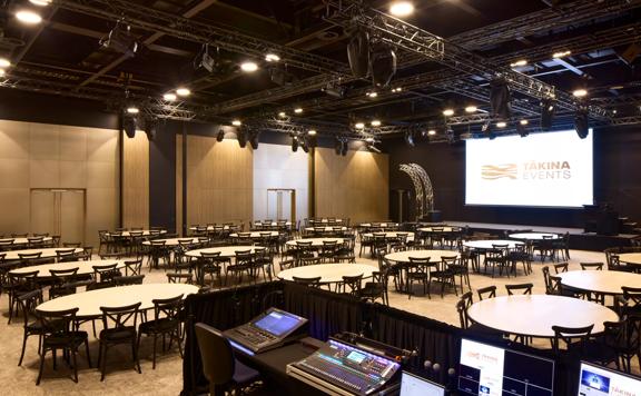 In an event space at Tākina, white tables fill the room with black chairs. An audio-visual sound desk is set up in the foreground, with a projector screen on the far wall.