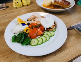 Plate of food on a wooden table featuring sliced cucumber, smoked salmon, greens, prawns, and poached eggs, with another plated meal visible in the background.