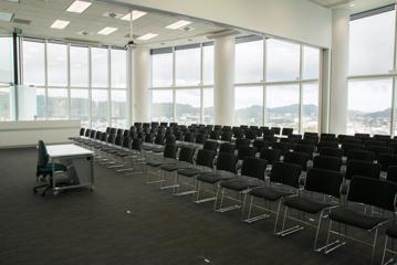 Large conference room with floor to ceiling windows and roughly 100 chairs facing towards camera.