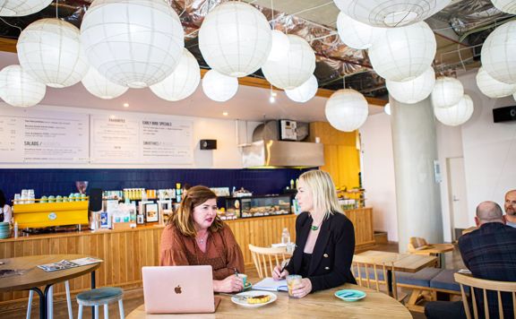 Two people sit at a round table in a café. They have a laptop, a couple coffees, a pastry on a plate and are taking notes while having a chat.