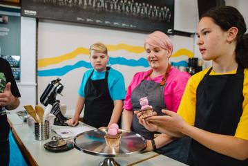 A film still from ‘Extreme Cake Sports’, a Wellington-based reality TV show. Three people are behind a counter, one holds a cupcake decorated with a pink pig made of icing.