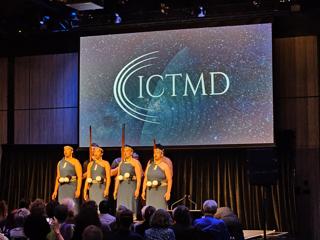 Six people on stage wearing traditional Māori clothing. The ICTMD logo is displayed on a projector screen behind them.