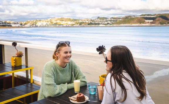 Two people sit and enjoy milkshakes and a dessert on the beach-side patio at Maranui café in Lyall Bay, Wellington.