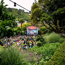 Looking through trees at the Botanical Gardensd at the stage and people sitting on the lawns at Gardens Magic 2023.