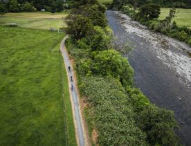 Birds eye view of three people cycling on a gravel path. On the left the path is lined with farmland and on the right a river.