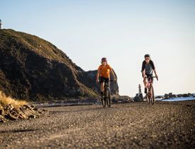 Two peple riding bikes on a gravel coastal path next to a large hill. Two lighthouses are visible in the distance, one on the coast line and one at the top of the hill.