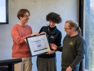 Three people standing in front of a concrete wall hold and admire a framed Rainbow Tick certification awarded to the Wellington Zoo.