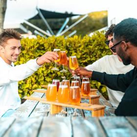 Three people sitting at a wooden picnic table on a patio at Brewtown in Upper Hutt, Wellington. They each have a flight of beers and are doing a cheers. 