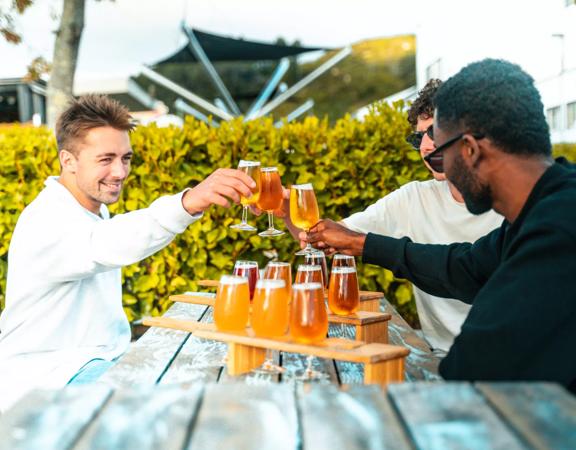 Three people sitting at a wooden picnic table on a patio at Brewtown in Upper Hutt, Wellington. They each have a flight of beers and are doing a cheers. 