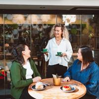 A smiling server brings a couple of coffees to two friends at a patio table.