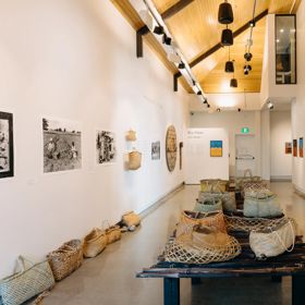 An exhibition room inside Toi MAHARA Gallery with woven baskets, photographs and other artworks on display.