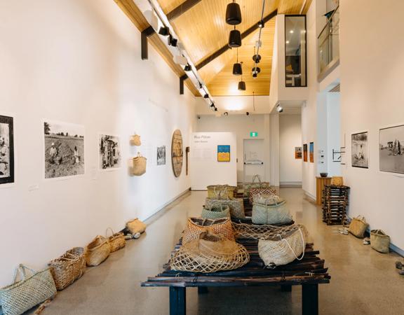 An exhibition room inside Toi MAHARA Gallery with woven baskets, photographs and other artworks on display. 