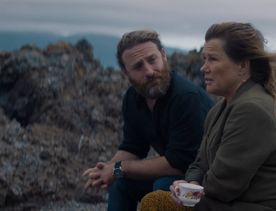 A production still from the series After the Party filmed in Wellington. Penny (Robyn Malcolm) is holding a teacup and sitting with Simon (played by Dean O'Gorman) on a rocky shoreline with a cloudy sky above.