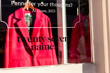 A bright pink blazer hangs in the window of a boutique clothing store called 'twenty-seven names'.