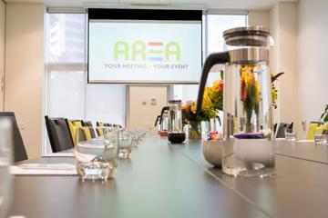 Table inside a conference room, with pitcher of water and glasses, Flowers on table and a projector screen in background depicting AREA's logo.