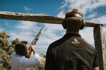 upwards angle of person shooting gun into air and person in foreground watching, both with earmuffs.