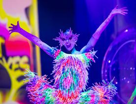 A model poses with raised arms, wearing a dress and sunglasses made of glowsticks.