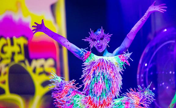 A model poses with raised arms, wearing a dress and sunglasses made of glowsticks.
